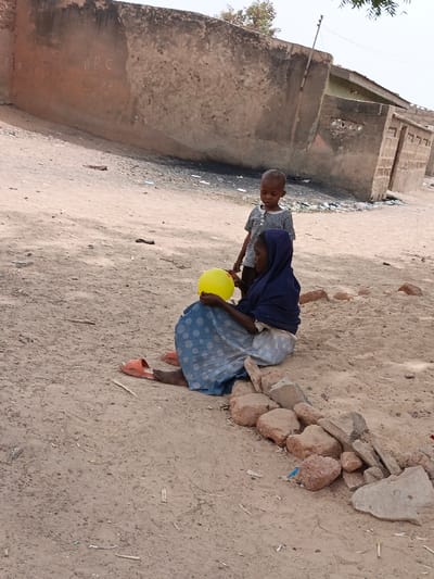 This image captures an outdoor scene in Zango, Nigeria. A seated adult female, wearing a dark blue head covering and a light blue polka-dotted skirt, holds a bright yellow spherical balloon. She sits on dry, light brown dirt ground, leaning against a low line of reddish-brown rocks. Her bare feet are visible next to a pair of orange slide sandals. Standing directly in front of her, a young child, approximately 3-5 years old, in a striped t-shirt, observes the balloon. In the background, a large, weathered light brown wall, possibly concrete or mud brick, exhibits graffiti including the letters "APC." Beyond this wall, single-story structures with small rectangular openings are visible. Scattered debris and litter, including white and dark fragments, are present on the ground near the base of the background structures. Sparse green tree foliage is visible against a light sky in the upper frame.