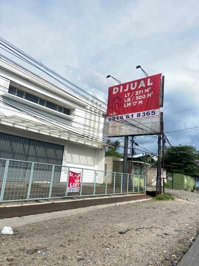 The image shows a white building with a large red sign that reads "DIJUAL" and displays the following information: "LT / 371 M2, LB / 500 M2, LM 17 M", and a phone number "0816 61 8365." The sign is mounted on a metal pole. In front of the building is a fence made of metal wire, a smaller red sign displaying "LUSTY" and the phone number "0816 61 8365" can be seen next to the fence. There are numerous electrical wires overhead and a narrow street in front of the fence. The coordinates -6.968, 107.616, suggest the location of the building is within the area of Kecamatan Dayeuhkolot, Bandung Regency, West Java, Indonesia. A small green building with a sign that reads "48" is partially visible in the background, along with some trees and a cloudy sky. The scene appears to be an urban or suburban setting. There are no people in the image.
