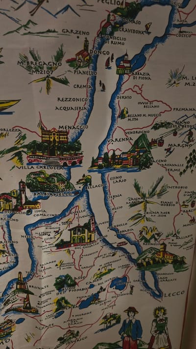 The image is a decorative, illustrated map of a lake and surrounding towns, possibly a souvenir or textile. It is not located in Vlaardingen, Netherlands. The map depicts a body of water with several towns and villages labeled with their names and some with altitudes (e.g., M. 2107). There are artistic renderings of buildings, churches, mountains, and boats on the water. In the lower right corner, there are illustrations of two people in traditional clothing. The style is colorful and somewhat cartoonish.