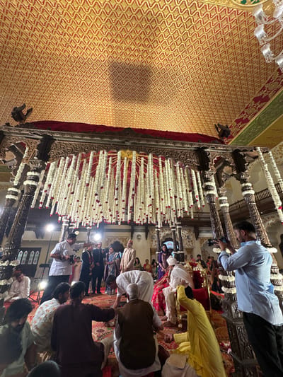 The image depicts an indoor wedding ceremony occurring within the Laxmi Vilas Palace in Vadodara, India. The scene is set in a grand hall beneath a high, ornate ceiling featuring a red and gold geometric patterned design. A substantial, intricately carved dark wooden structure, identified as a mandap, occupies the central foreground. This structure is heavily adorned with numerous vertical strings of white flowers, accented with sporadic red elements, suspended from a circular fixture beneath its canopy.
At the center, a couple, presumably the bride and groom, is seated on an ornate red chair. The groom wears a white turban and traditional attire. Surrounding the couple, approximately twenty individuals are visible. Several attendees are seated directly on the patterned reddish-orange carpeted floor in the foreground, participating in a ritual; some are kneeling or bending forward over small metallic vessels placed on the carpet.
Two professional photographers/videographers are actively documenting the event. One individual in a white shirt is positioned on the left, holding a camera with an attached flash unit, pointed towards the couple. Another individual in a light blue shirt is positioned on the right, operating a video camera, also directed towards the central figures. A light stand with a flash is visible on the left side, providing additional illumination. Other guests stand in the mid-ground and background, observing the ceremony. Balconies with carved wooden railings are visible in the upper background, indicating a multi-story interior space.