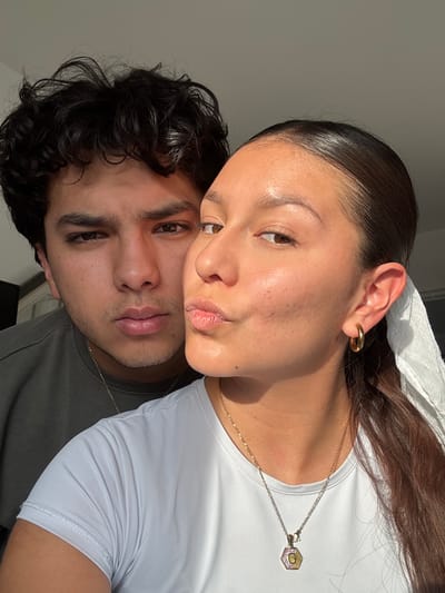 A close-up portrait captures a young adult couple in what appears to be an intimate and relaxed pose, with the image rotated 90 degrees clockwise. The female subject, with dark, neatly slicked-back hair and visible baby hairs, gently rests her head against the male subject. Both look towards the camera with calm, slightly parted lips.

The female is wearing a white, athletic-style top, showcasing a smooth neckline. She accessorizes with a golden ear cuff on her right ear. A delicate gold chain necklace hangs around her neck, featuring a distinctive hexagonal pendant with a visible initial, likely a 'G', inset against a pearlescent background. Her skin appears naturally luminous under the bright lighting.

The male subject, positioned beneath the female, has dark, curly hair and a subtle amount of stubble on his face. He is wearing a dark grey or greenish t-shirt, and a thin gold chain is also visible around his neck.

The scene is illuminated by strong, natural light, likely sunlight, casting a warm glow and creating highlights on their skin and hair, suggesting it is daytime. The background is minimal, featuring a plain, light-colored wall on the left and a darker, indistinct area on the right, indicating an indoor setting. The overall mood conveyed is one of quiet affection and serenity. No specific location details are visible within the frame, but the provided context places it in San Francisco Ocotlán (Ocotlán), Mexico.