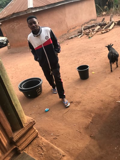 A young adult male is centrally positioned outdoors in Ogodo, Nigeria, standing on reddish-brown dirt. He wears a color-block track jacket (white, red, black), black track pants with red stripes, and light blue and white flip-flops. His hands are in his pockets, and he smiles slightly while looking directly at the camera. A gold-colored bracelet is visible on his right wrist. To his left, two black plastic buckets are nested, with the word "ELF" visible on the upper bucket. A single black plastic bucket is positioned to his right. A black goat with visible horns stands towards the right, facing away from the camera. In the background, a one-story building with a light brown wall and a corrugated metal roof is visible, with a pile of cut tree branches and logs to its right. A silver vehicle is partially visible on the far left. Scattered on the dirt ground are a crumpled light blue wrapper, a small yellow piece of debris, and other small natural fragments. A textured, reddish-brown architectural element occupies the lower left foreground.