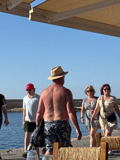 This image captures a scene on a sunny day in Chania, Greece, likely near the waterfront. In the foreground, a shirtless man wearing a straw hat and patterned swim trunks stands with his back to the camera. To his left, another man in a light-colored polo shirt and dark shorts, wearing a red baseball cap and sunglasses, is walking away. Further to the left, a portion of a third person is visible, wearing a black t-shirt. In the mid-ground, two women are walking towards the foreground. The woman on the left wears a patterned dress and sunglasses, and the woman on the right wears a floral print dress, sunglasses, and a cross-body bag. The background consists of calm blue water, a stone wall, and a clear blue sky, with a portion of a canvas canopy visible at the top of the frame. The overall setting suggests a casual outdoor environment, possibly a promenade or a beachside area.