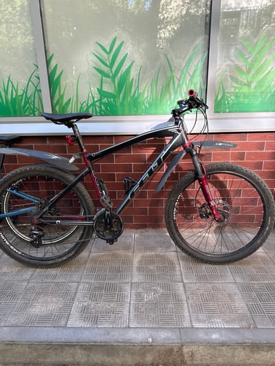 A black and red mountain bike is parked on a tiled sidewalk in front of a brick wall and a window with a green grass design in Bykovo, Russia. The bike has mudguards on both wheels and a red rear light. The front wheel is a disc brake wheel. The bike is a Fuji model.