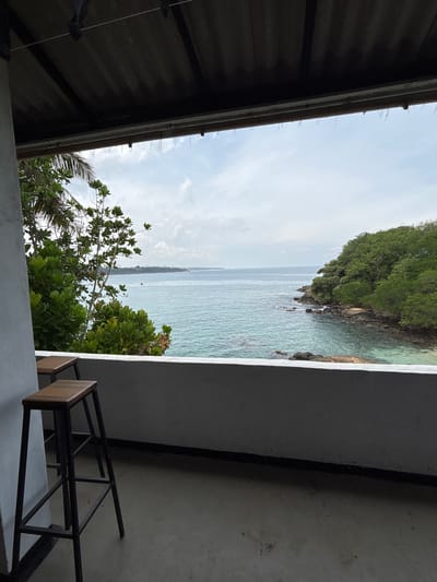 The image depicts a view from a balcony or window in Dikwella, Sri Lanka. The scene is framed by the architecture of the building, including a dark roof on the left, a section of wall in the foreground, and a vertical wall on the right with stairs. Beyond the structure, the view features a body of water, possibly the ocean, with a landmass covered in lush green vegetation extending into the water on the left side of the frame. The sky is visible above, with a mix of clouds and clear sky. In the foreground, vegetation including palm and leafy trees partially obscures the view of the ocean.