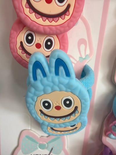 The image displays a close-up of several cartoonish, fuzzy monster-themed hair accessories, likely hair ties or bracelets, against a plain, light-colored background. The central and most prominent item is a light blue fuzzy monster with large, round eyes, a wide grin with jagged teeth, and blue bunny-like ears. The monster's face is a light tan color. Behind it, slightly out of focus, are two pink fuzzy monsters with similar features. Below the blue monster, a pink and light blue polka-dotted bow is visible. The overall aesthetic is playful and childlike. The image does not contain any information or visual cues that would allow for the identification of a specific location in the city of Ravno pole, Bulgaria.