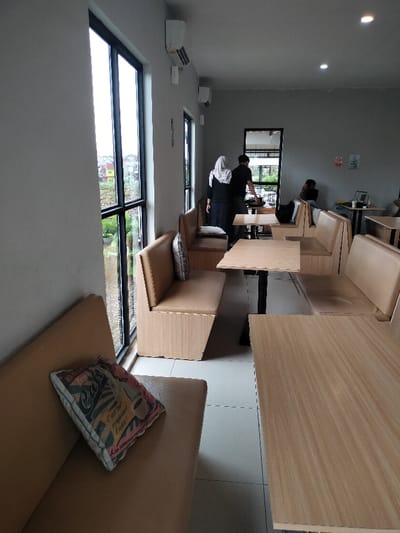 The image depicts the interior of a modern cafe or restaurant in Dramaga, Indonesia, characterized by a minimalist aesthetic. The walls are light gray, and the floor is tiled in white. Large, floor-toceiling windows line the left side, offering a view of a green, outdoor landscape with trees and distant residential buildings, including a distinct yellow house.
The cafe is furnished with booth-style seating. Light wood benches with beige upholstered cushions are arranged around light wooden tables with dark metal bases. In the foreground, one cushion features a decorative print with French text, including "Cafe" and "C'est tout simplement Expres," along with an illustration of a coffee cup.
Several individuals are present in the cafe. In the mid-ground, two people are standing near a counter or table: one wearing a white hijab and dark clothing, and another in a dark t-shirt. They appear to be interacting or attending to something. Further back on the right, a person is seated at a booth with a laptop on the table.
The ceiling features recessed lighting, and multiple white wall-mounted air conditioning units are visible on the gray walls. A "No Smoking" sign is also visible on the far wall. The natural light flooding through the large windows suggests it is daytime. The overall atmosphere appears calm, with a few patrons and staff in the space.