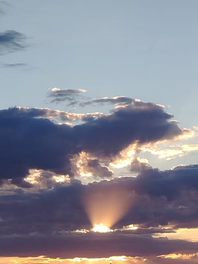 A vertical, medium shot captures a dramatic sunset over Baikonur, Kazakhstan. The sky is a pale blue, fading to a warmer yellow-orange at the horizon, where the sun's rays are just visible through a bank of dark, textured clouds. These clouds are illuminated from behind, creating a halo effect and casting a warm glow. In the center of the frame, a distinct sunbeam radiates outwards from behind a thick cloud mass, creating a crepuscular ray effect. The clouds themselves vary in color from deep indigo to lighter shades of grey and purple, with the edges highlighted by the setting sun. The lower portion of the image shows a sliver of land, possibly a building or structures, silhouetted against the vibrant horizon. No people or animals are visible.