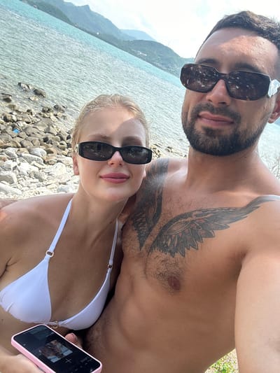 A man and a woman are taking a selfie on a rocky beach. The man is shirtless, revealing a large tattoo on his chest. The woman is wearing a white bikini top. The ocean is visible behind them, with mountains in the distance. The man is wearing dark sunglasses, and the woman is wearing black sunglasses. The woman is holding a pink smartphone. The location is the city of Ninh Ích, Vietnam.