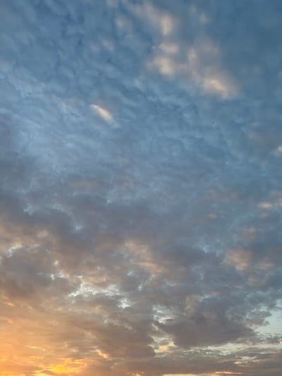 The image shows a sky with clouds during sunset or sunrise. The lower part of the sky is illuminated with warm orange and yellow hues, characteristic of the setting or rising sun. As the eye moves upwards, the colors transition to cooler blues and grays, with scattered white and gray clouds forming patterns across the sky. There are no discernible buildings, landmarks, or natural features visible that would indicate a specific location within Poing, Germany. The image is a close-up view of the atmospheric conditions, with no human presence or activity depicted.