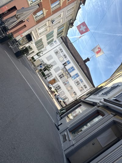 The image captures a sunlit street scene in Liestal, Switzerland, presented with an upward and tilted perspective along the thoroughfare. The setting is an urban environment characterized by traditional European architecture.
Prominently displayed in the upper part of the frame are two flags suspended across the street: the national red Swiss flag with a white cross, and a white municipal flag bearing a red, stylized crozier, representing Liestal. The sky is bright blue with scattered white clouds and several visible aircraft contrails, indicating a clear day.
The street is lined with multi-story buildings exhibiting various facades, including light peach, beige, white, and grey. Most windows are adorned with shutters, either closed or partially open, in muted green or grey tones. One central white building features a decorative balcony with a circular emblem or clock face visible beneath it. On the left, a building with a peach-colored facade hosts outdoor seating, including a large, beige umbrella and dark chairs, and a visible sign that reads "LUTHY".
The asphalt road surface is visible in the foreground and extends into the distance. A white car is parked further down the street, near which a single person is standing, and a silver van is also visible. Street furniture such as planters with small trees and a wooden bench add to the urban ambiance. The bright sunlight casts clear shadows, suggesting it is either morning or late afternoon.