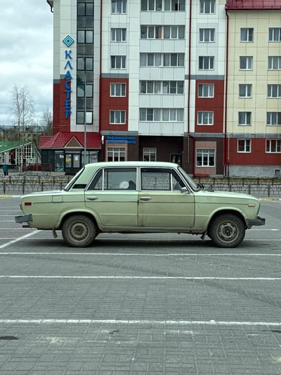 The image depicts a light green sedan parked in a gray brick parking lot. The car is adjacent to a multi-story building with a sign in Cyrillic script, "КВАРТЕЛ." The building has multiple windows and is colored in sections of cream, white, and red. Part of another building with a red roof is also visible, located across from the larger building. A metal barrier or fence is situated near the smaller building. Bare trees are visible in the distance under a cloudy sky. The location is identifiable as the city of Beloyarsky, Russia, due to the Cyrillic signage.