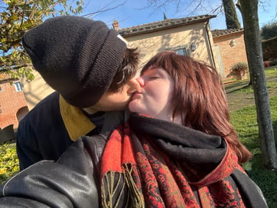 The image displays two individuals engaged in a kiss outdoors on a sunny day, situated within a garden or yard in Montepulciano, Italy. The subject on the left wears a black knit beanie, a dark jacket with a yellow collar, and has visible facial stubble; their profile shows lips pressed against the other subject's. The subject on the right has shoulder-length reddish-brown hair with bangs, and wears a black leather jacket over a black garment, accessorized with a red, black, and gold patterned scarf featuring tassels; their eyes are closed and lips pursed. The background features traditional Tuscan architecture, including a light-colored building with a terracotta tiled roof, two windows, and wall sconces, and a partial reddish-brown brick structure with an archway. The ground is covered in green grass, with several trees, including a prominent tree trunk on the right, and sparse foliage visible against a clear blue sky.