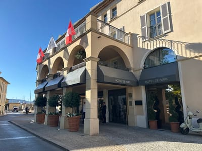 The image shows the facade of the Hôtel de Paris in Saint-Tropez, France. The hotel has a light-colored stucco exterior with arched entrances and balconies. Several flags are visible, including three Swiss flags and one white flag with text. Two large planters with trees and white flowers flank the main entrance, which features glass doors with the hotel's name displayed above. A light-colored scooter is parked to the right of the entrance. In the background, a street is visible with cars and buildings. The sky is clear and blue.