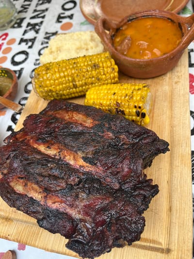 The image shows a wooden platter with a hearty meal. Featured are a rack of ribs with a dark, caramelized crust, two ears of grilled corn, and a small terracotta pot filled with a stew. Beside the corn is a small portion of white mashed or puréed food. The platter rests on a surface covered with a patterned tablecloth. The scene evokes a sense of rustic dining, possibly outdoors. The location given is San Francisco Totimehuacán, Mexico.