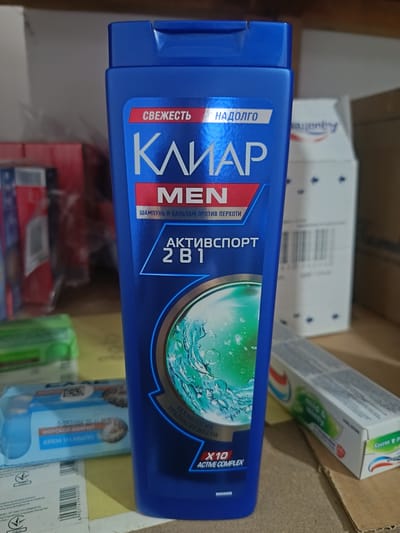 This image shows a close-up of a blue bottle of "Clear" brand shampoo for men, with Russian text on the label. The bottle is positioned on a surface, possibly a shelf or table, with other items visible in the background, including a box of toothpaste and what appear to be other product boxes or packaging. The lighting is direct, highlighting the glossy surface of the shampoo bottle. There is no visible indication of the city of Berdiansk, Ukraine, or any environmental details that would place the scene in a specific urban location. The focus is entirely on the product and its immediate surroundings.