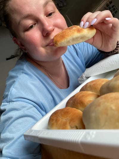 A close-up, over-the-shoulder shot shows a person in a light blue t-shirt about to eat a baked pastry. The pastry is golden brown and oval-shaped. The person's hand, with light purple nail polish, is holding the pastry to their mouth. In the foreground, a white tray holds several similar pastries. The background is blurred, but appears to be an indoor setting. The image does not provide any specific location details within the city of Балдейка, Russia.