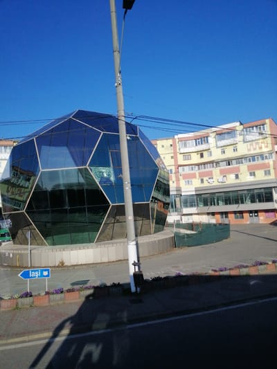The image shows a geodesic dome-shaped building with a glass facade, reflecting the surrounding environment. The building is located in Roman, Romania, as indicated by a blue directional sign that reads "Iași". The dome is a prominent structure, situated in an urban setting with a multi-story building in the background that has a yellow and pink facade. Several windows and balconies are visible on this building, with some laundry hanging out to dry. In front of the geodesic dome, a paved area is visible, with flowerbeds lining the edge. A lamppost is present in the foreground, casting a shadow onto the paved surface. The sky is clear and blue.