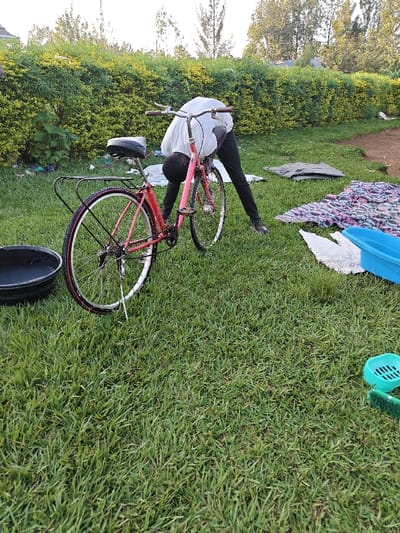 In an outdoor grassy area within Ang'orom ward, Kenya, a male individual is depicted bent over a pink-framed bicycle. He wears a white long-sleeved shirt and dark trousers, with his head positioned near the bicycle's frame as if performing maintenance or inspection. The bicycle, equipped with a black seat, silver handlebars, and a rear rack, stands upright. To the left of the bicycle's rear wheel, a black plastic basin rests on the grass. To the right, various items are spread on the grass, including a patterned blanket (blue, pink, white), a grey garment, and a light blue rectangular plastic tub. In the lower right foreground, a green plastic soap dish and a green scrubbing brush are visible. A dense green hedge forms the background, with trees and a bright sky beyond. The ground consists of well-maintained green grass.