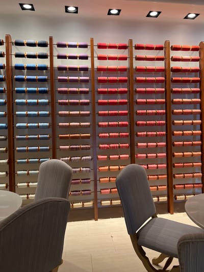 The image shows a retail display of spools of thread or yarn arranged on wooden racks against a plain white wall. The spools are organized in horizontal rows, with each row containing multiple spools. The colors of the spools vary from dark blue and purple to various shades of pink, red, and orange, as well as some neutral tones like beige and white. There are three prominent wooden racks visible, each holding multiple rows of spools. In the foreground, two grey upholstered chairs are partially visible, positioned in front of the display. A round, light-colored table is also partially visible to the left. The lighting appears to be from overhead spotlights. The scene suggests a fabric store or a similar retail establishment selling craft supplies. The specific location in Port De Pollença, Spain is not discernible from the image itself.