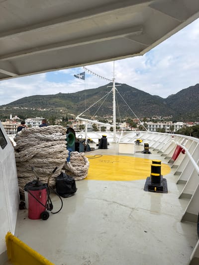 On the deck of a ship near the Municipal Unit of Methana, Greece, a large coil of rope rests near a red and black wheeled object and a black plastic bag. Mountains and buildings of Methana are visible in the background. The deck is painted with white and yellow markings, and black mooring bollards are visible. A Greek flag flies from a mast on the ship.