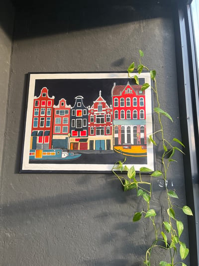 A framed painting of Dutch-style canal houses with a dark background hangs on a textured gray wall. A pothos plant with green and yellow variegated leaves is draped over the top right corner of the painting and trails down the wall. The painting is bordered by a white mat and a dark frame. To the right of the painting, a dark metal window frame is visible. Sunlight casts a shadow on the upper left portion of the wall. The setting appears to be indoors, possibly a cafe or a home, in the city of Prigorodny, Russia.
