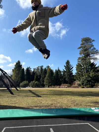 A young person is captured mid-air against a bright blue sky with scattered clouds. They wear a tan fleece jacket, grey sweatpants, black shoes, a yellow beanie, and red gloves. Their arms and legs are outstretched, suggesting a jump or fall. The backdrop includes green trees, a grassy area, and part of what appears to be a trampoline with a green safety pad. A metal structure is visible in the lower left. The location is the city of Stapriņi, Latvia.