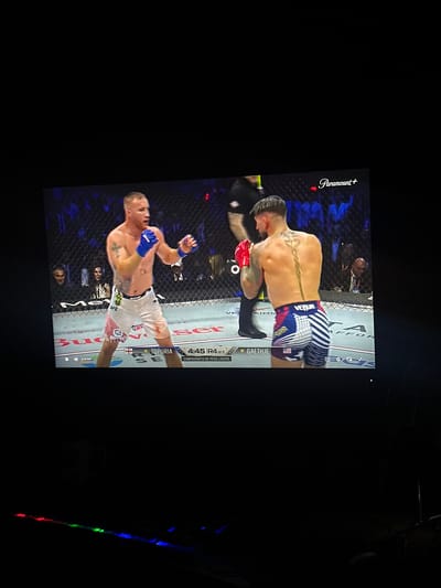 The image shows a screen displaying a UFC fight. Two male fighters are in the center of the octagon, facing each other. The fighter on the left is wearing white shorts and blue gloves, with tattoos on his chest and arms. The fighter on the right is wearing red gloves and patterned shorts with red, white, and blue stripes, and has a distinctive tattoo running down his back. The Paramount+ logo is visible in the top right corner of the screen. Below the fighters, text indicates "TOPURIA" and "GAETHJE", along with "4:45 R4 OF 5" and "CAMPEONATO DE PESO LIGERO". The background shows a blurred crowd watching the event. The lower part of the image is dark, with a glowing keyboard visible, suggesting the screen is a television or monitor in a room. There is no indication of this scene being located in Perímetro Urbano Barranquilla, Colombia.