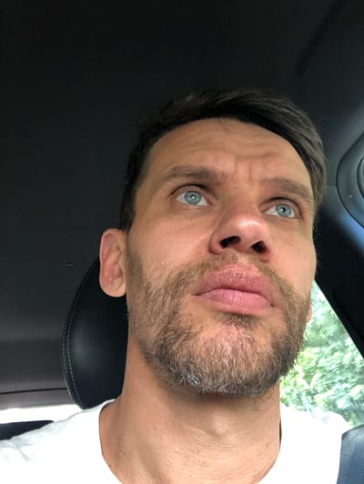 A close-up, low-angle, outdoor selfie of a man with blue eyes, a light brown beard, and short brown hair, looking upwards. He is wearing a white t-shirt and is seated in the driver's seat of a car, with the headrest visible behind him. The background shows a blurred view of green foliage, suggesting a natural setting. The man's facial expression appears thoughtful or questioning. The image was taken in Seltso, Russia.