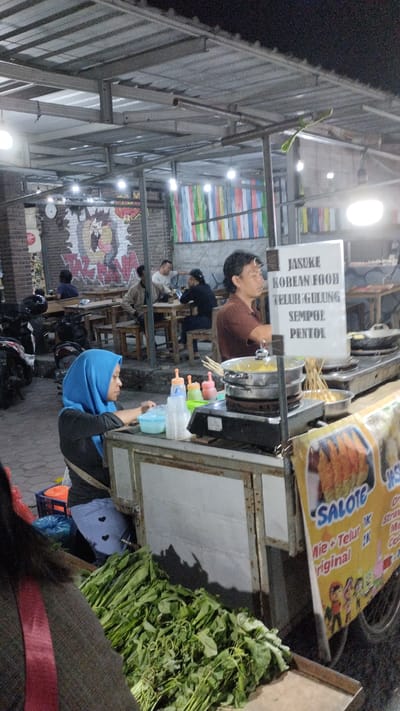 This media file captures a lively street food scene at night, likely in Sukodono, Indonesia. The central focus is a food stall with a metal roof and frame, illuminated by overhead lights. A woman wearing a blue hijab sits behind the counter, engaged in preparing food. In front of her are various condiments and utensils. To her right, a man in a brown shirt stands, seemingly attending to the food being cooked.
The stall displays a sign with text that appears to list food items, including "JASUKE," "KOREAN FOOD," "TELUR GULUNG," "SEMPOL," and "PENTOL." Another sign on the side of the stall reads "SALOTE" and lists prices for items like "Mie + Telur" and "Original." Piles of fresh green vegetables are neatly arranged in front of the stall, suggesting the availability of fresh ingredients.
In the background, several people are seated at wooden tables, enjoying their meals and conversing, indicating a busy dining area. The setting appears to be an outdoor market or a street lined with food vendors, with motorcycles parked nearby. The overall atmosphere suggests a casual and bustling evening scene, characteristic of Indonesian night markets. The time of day is clearly night, as evidenced by the artificial lighting and the dark sky. There are no strong indicators of weather, but the presence of overhead covering suggests it might be a covered area to protect from elements.