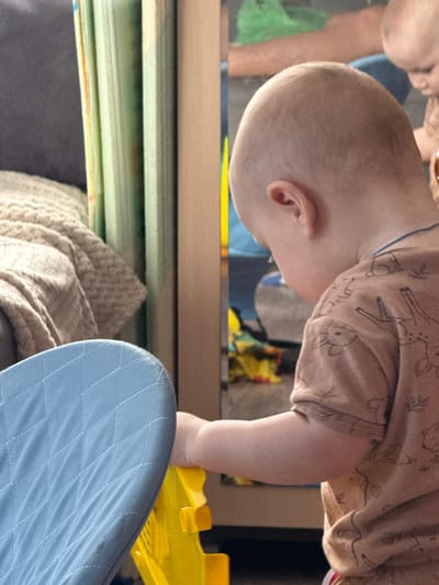A baby in a patterned shirt is reaching towards a yellow toy near a blue quilted seat. In the reflection of a surface above, a person's face is partially visible. Below, there are stacked objects and a beige knitted item on a gray surface. The location is Кипчак, Russia.
