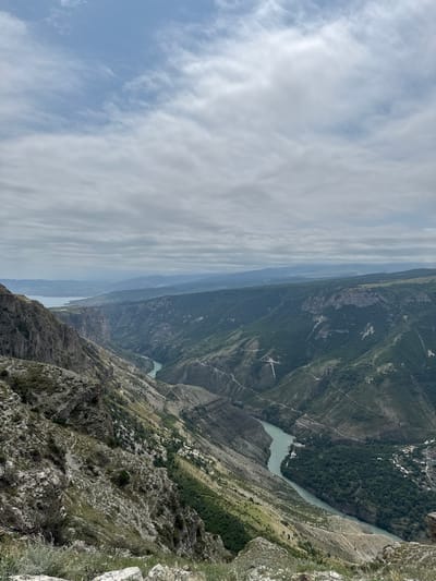 The image captures a panoramic view of a vast, deep canyon with a winding river flowing through it. The canyon walls are steep and rugged, with layers of rock formations visible. Vegetation, including trees and shrubs, covers the slopes, particularly in the lower sections. A road or path can be seen traversing the canyon's side in several places. In the distance, the river widens into a body of water, possibly a lake or reservoir, with hills and mountains stretching to the horizon under a cloudy sky. On the right side of the canyon, a small settlement with buildings and a bridge crossing the river is visible. The perspective suggests the photo was taken from a high vantage point, overlooking the immense natural landscape. The scene appears to be the Sulak Canyon, a prominent natural landmark located near the city of Dubki, Russia. There are no people or animals visible in the image.