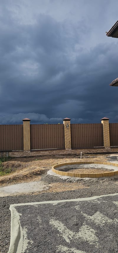 The media file displays an outdoor scene under a cloudy, dark sky, suggesting overcast weather and possibly impending rain. The perspective is from a low angle, looking upwards, with the image rotated 90 degrees counterclockwise.
The primary subjects are a brown metal fence with decorative brick pillars at intervals. These pillars are light yellow and topped with darker brown caps. Behind the fence, a section of a building's roofline is visible, featuring a dark brown overhang. To the right of the fence, the ground is covered in a mixture of gravel and dry, yellowish grass. A circular structure made of light yellow bricks, possibly a garden bed or a decorative feature, is partially visible on the gravel. A white pipe protrudes from the ground near the fence.
The overall impression is that of a residential or backyard setting in a rural or suburban area. The dark clouds create a somewhat somber mood. The time of day appears to be daytime, given the illumination, but the cloudy conditions obscure direct sunlight. There is no visible text in the image.