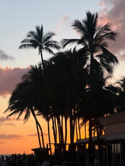 The image shows a sunset scene with palm trees in the foreground. The sky is a gradient of colors, ranging from light orange near the horizon to pink and purple at the top. The palm trees are silhouetted against the sky.

In the lower right corner, there is a building, and there are people.

Based on the presence of palm trees, the sunset over the ocean, and the style of the building, it's plausible to say that the photo may be from Honolulu, United States of America.