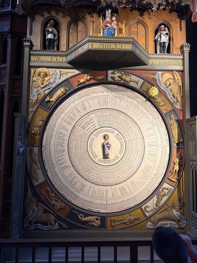 The image shows the astronomical clock in Lund Cathedral, Sweden. The clock is a large, circular mechanism adorned with figures and inscriptions. At the top, above the clock face, are statues of religious figures, including a Madonna and child, and figures in armor holding trumpets. The clock face itself is a complex arrangement of concentric circles with text and numerical data, suggesting it displays astronomical and calendrical information. Around the perimeter of the clock face are depictions of zodiac signs and carved figures representing months or celestial beings, with labels such as "JANUARI," "FEBRUARI," "MARS," "APRILL," "SEPTEMBER," "OKTOBER," and "NOVEMBER." Golden statues of angels and mythological creatures are also integrated into the clock's design. The overall impression is one of intricate craftsmanship and historical significance, characteristic of medieval or early Renaissance astronomical devices. The clock is housed within the dark, ornate interior of Lund Cathedral, with visible wooden architectural elements and decorative carvings.