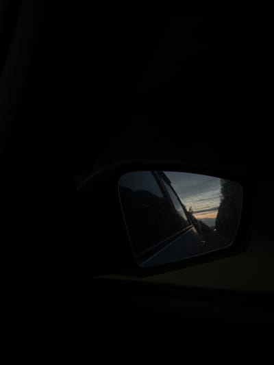 This is a close-up, dark, outdoor shot of a car's side mirror, reflecting a cloudy sunset over a road lined with trees. The mirror is framed by a black car interior, and the reflection shows a road curving into the distance, with a hint of orange and pink in the sky. The image is undated and the location in Okulov, Russia is not visually apparent from the content.