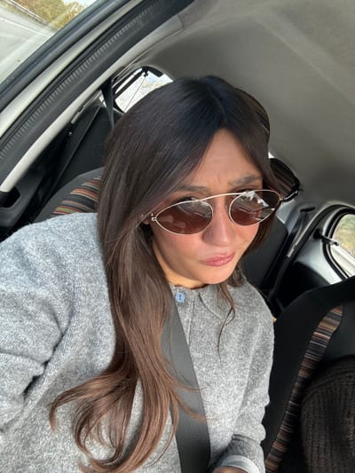 The image captures a selfie taken inside a vehicle, depicting an adult female in what is presumed to be Le Cannet-des-Maures, France. The subject, positioned in the passenger seat, is visible from the chest upwards, looking downward and slightly to her right with pursed lips. She has long, dark brown hair that falls over her left shoulder and chest. She wears gold-framed, oval-shaped sunglasses with dark brown reflective lenses. Her attire is a light gray, textured sweater or cardigan, with a single light blue button visible near her right collarbone. A dark-colored seatbelt crosses diagonally from her left shoulder across her torso. The car's interior features a light-colored roof lining, and the seat behind her (driver's side) has a headrest with multi-colored stripes (brown, orange, black, beige). Through the window on the upper left, a blurred outdoor environment with a road and green-brown foliage is visible, suggesting movement. No identifying landmarks or specific street signs are present to visually confirm the exact location within Le Cannet-des-Maures.