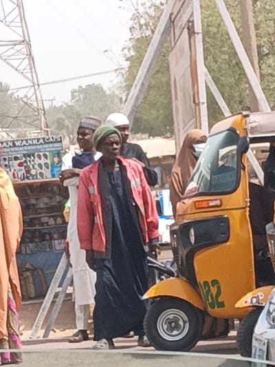 This image depicts an outdoor urban scene in Hotoro South, Nigeria. In the foreground, an older individual wearing a dark blue tunic, a red long-sleeved jacket, and a green knitted cap stands slightly left of center, looking towards the right. To their left, a younger male in a white long-sleeved garment and a patterned dark blue cap is visible. Further right, partially obscured, a male wearing a dark garment and a white skullcap stands. On the far right, a yellow three-wheeled auto-rickshaw, marked with "32" in green, is parked or waiting; a person in a light brown hijab and a light blue face mask is partially visible behind its front section. In the far left foreground, a person in a light brown garment with a colorful hem is partially cropped. The background features a vendor's display sign reading "DAN WANKA CAP" with various cap designs. A large metal lattice tower stands in the distant left background, while other vertical metal structures are present further right. Sparse trees and shrubs are visible against a bright, clear sky. The ground in the immediate foreground appears paved, transitioning to a dirt or unpaved area in the background. People are standing or moving in what appears to be a street or market environment.
