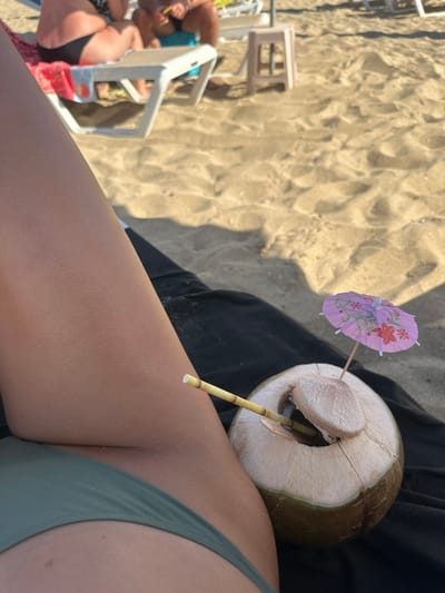 The image depicts a close-up view of a person's leg and a coconut drink on a sandy beach. The person is wearing a green bikini bottom. The coconut is partially open with a straw and a small pink umbrella inserted into it. In the background, blurred figures of people and beach furniture are visible, suggesting a public beach setting. The sand is light brown and appears undisturbed, indicating it is a typical beach environment. The lighting suggests it is daytime, likely sunny, casting shadows on the sand. The context of Protaras, Cyprus, implies a coastal Mediterranean location known for its beaches.