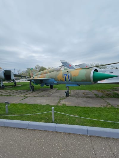 The image shows a MiG-21 fighter jet displayed outdoors, likely at a museum. The aircraft has a camouflage paint scheme and the number "17" on its side. It is positioned on concrete slabs surrounded by grass, with a gray curb separating the display area from an asphalt surface. In the background, other aircraft are visible, indicating this is part of an aviation exhibit. The sky is overcast. The location is Arkhangel’skoye, Russia.