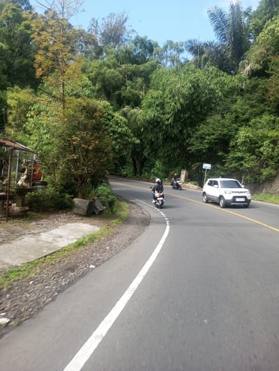 The image depicts a curved road in Khe Luang, Laos, surrounded by dense green vegetation, including various trees and possibly bamboo. The road has a white dividing line.
A white car and two motorcycles are on the road. The car is positioned in the oncoming lane. The motorcycles are driving in the same direction as the viewpoint.
To the left of the road, there is a concrete walkway alongside what appears to be a small shop or establishment with plants and decorative objects. The sky is partly clear with a slight blue tint.