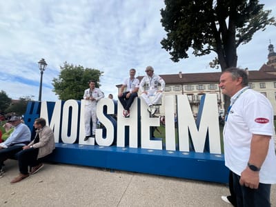 This image depicts an outdoor public gathering in Molsheim, France, under an overcast sky. The central feature is a large, three-dimensional sign spelling "#MOLSHEIM" in white uppercase letters on a blue base, positioned on a paved surface, likely in a public square or park.

Three men are situated on the sign. From the left, a man in a white full-body racing suit with patches and a gold medal around his neck stands on the letter 'L'. To his right, a man wearing a light-colored polo shirt, dark trousers, and red and white sneakers sits on the letter 'S'. Further right, a third man in a white full-body racing suit, a white beanie, and sunglasses sits on the second letter 'M'.

In the right foreground, a man partially visible from the chest up, with a mustache and short brown hair, is shown looking towards the left. He wears a white short-sleeved shirt with a red "BUGATTI" logo on the left sleeve and a blue event lanyard around his neck. A dark-colored watch is on his left wrist. Several other individuals are partially visible in the left mid-ground, some seated on a low green surface.

The background reveals a multi-story, historic European-style building with numerous windows and a reddish roof, topped by a visible bell or clock tower structure. Mature green trees are present behind the sign, and an ornate black lamppost stands on the far left.