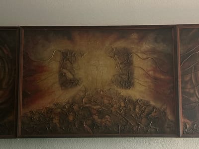 The image displays a multi-panel abstract artwork mounted on a light-colored, textured wall indoors. The central panel is a rectangular piece featuring a highly textured surface composed of crumpled material and linear elements. Its color palette primarily consists of earthy browns, deep reds, and ochres, with a lighter, possibly illuminated, central area and darker, rougher forms below and to the sides. Wavy, thin lines, appearing metallic, traverse sections of the artwork. The entire composition, including partial views of adjacent panels on the left and right, is enclosed within a dark, likely wooden frame. Above the artwork, a flat, light-colored ceiling is visible. No individuals are present, and no actions or interactions are depicted. The image provides no visual information to identify its physical location as being in the city of Bogdanci, North Macedonia; the artwork itself is abstract and does not represent a specific geographical scene.