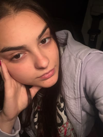 A young woman is depicted in a close-up, tilted selfie. She has fair skin, dark brown hair, and brown eyes. Her face is partially supported by her left hand. She is wearing a light purple hoodie with a visible silver zipper. Behind her is a patterned fabric with white, red, and black designs. The overall lighting is dim, and the background is dark. There is no indication of the location being Elenovo, Bulgaria.