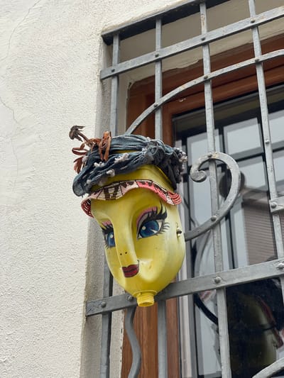 A stylized human face, crafted from an inverted yellow plastic container, is suspended from an ornate dark grey metal security grate on a building's exterior in Buñol, Spain. The face features painted blue eyes with prominent black eyelashes and pink eyeshadow, red lips, and two black dots for nostrils, with the container's spout forming the chin. The upper part of the 'head' is adorned with a dark grey fabric containing small white tufts, a band of pink and white beads across the forehead, and several dried brown plant elements protruding from the top. A single light-colored bead hangs as an earring from a small hole on the right side of the face. The metal grate, secured with rivets, covers a wooden-framed window whose glass reflects ambient light. To the left of the window, the building's facade is a textured, light-colored stucco wall with minor surface cracks.