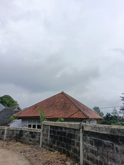 This image displays a static outdoor scene in Kabiufa, Papua New Guinea, under a heavily overcast sky composed of light grey clouds. In the middle ground, a dark grey concrete block wall with a lighter concrete coping extends horizontally across the frame, exhibiting signs of weathering and discoloration. A dirt or gravel path with sparse dry foliage is visible in the immediate foreground, to the left of the wall. Behind the wall, a house with a distinct reddish-orange terracotta tiled hip roof and light-colored walls featuring multiple horizontal windows is centrally positioned. A large green broad-leafed plant grows in front of this house. To the left, a second building with a dark grey tiled roof is partially visible. Green trees and foliage are present in the background on both sides. A single strand of barbed wire is discernible in the upper right portion of the frame. No individuals, animals, or active subjects are depicted within the scene.