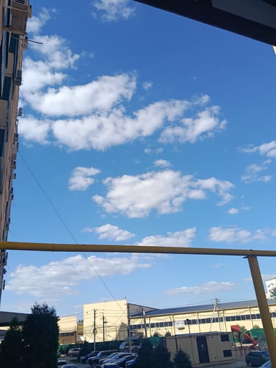 The image depicts a street scene in Yablonovskiy, Russia, on a clear, sunny day. The sky is a vibrant blue, dotted with scattered white clouds. In the foreground, a thick yellow pipe, possibly a gas pipe, runs horizontally across the frame, supported by vertical poles. To the left, the side of a multi-story building is visible, with numerous air conditioning units mounted on its exterior. Below the building and the yellow pipe, a row of parked cars is visible, including sedans and SUVs. Beyond the cars, several low-rise buildings with light-colored facades line the street, some appearing to be commercial or industrial structures. Green trees and shrubs are interspersed throughout the scene, providing a touch of nature amidst the urban environment. A playground with red structures is also partially visible in the background. The overall impression is one of a typical urban residential or commercial area.