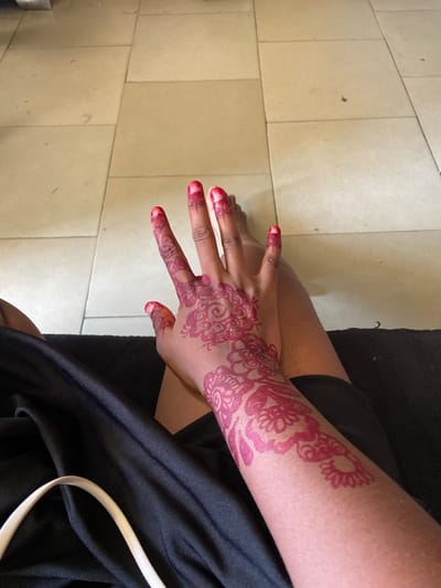 An aerial shot depicts a person sitting with their legs crossed. The person has a dark skin tone, a black shirt, and their left arm and hand are adorned with a detailed henna tattoo design in red. The henna design extends from the forearm to the hand, including designs on the fingers and fingertips. The person's hand and foot are partially visible. The background is tiled flooring with an off-white tone. The location is unknown.