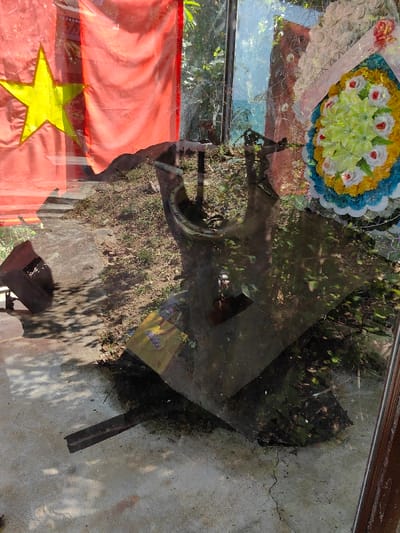 A display case exhibits war remnants alongside a Vietnamese flag. Inside, weathered metal fragments, possibly from a downed aircraft, rest on a concrete surface. Reflections on the glass show outdoor foliage and a large floral wreath, creating a layered visual effect. The setting evokes a somber, memorial atmosphere, contrasting the patriotic symbol with tangible artifacts of conflict.