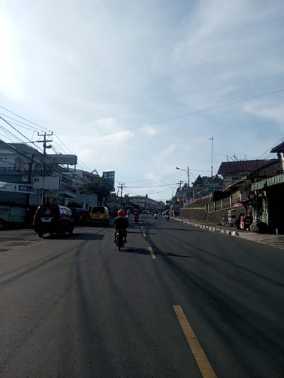 The image depicts a street scene in what appears to be a town or city. A two-lane road dominates the foreground, with a single yellow line indicating the lanes. A motorcycle, ridden by a person wearing an orange helmet, is positioned in the center lane of the road, moving away from the camera. Further ahead, there are more motorcycles and a few cars visible.
Buildings line either side of the road. On the left, there are multi-story buildings with signs, including one that reads "RYA INDAH" and another that mentions "3GB 11 ribu," suggesting a telecommunications advertisement. Vehicles, including a dark-colored minivan and a yellow car, are parked on the side of the road. Power lines crisscross overhead, adding to the urban landscape.
On the right side of the road, there are buildings and a slightly raised sidewalk with a low wall or embankment. A motorcycle is parked near a building. In the distance, the road appears to lead uphill, with more buildings and structures visible. The sky is partly cloudy, and the lighting suggests it is daytime. The overall impression is of a busy street in a developing urban area, possibly in Southeast Asia.